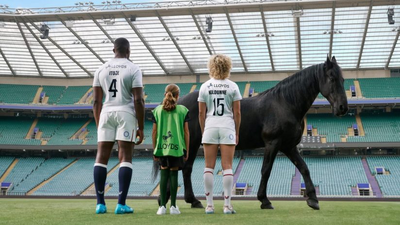 Maro Itoje, Lark Davies, and a child stand with a black horse on a rugby pitch, facing empty stadium seats.