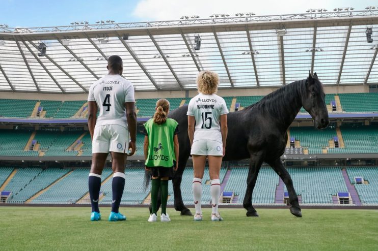 Maro Itoje, Lark Davies, and a child stand with a black horse on a rugby pitch, facing empty stadium seats.
